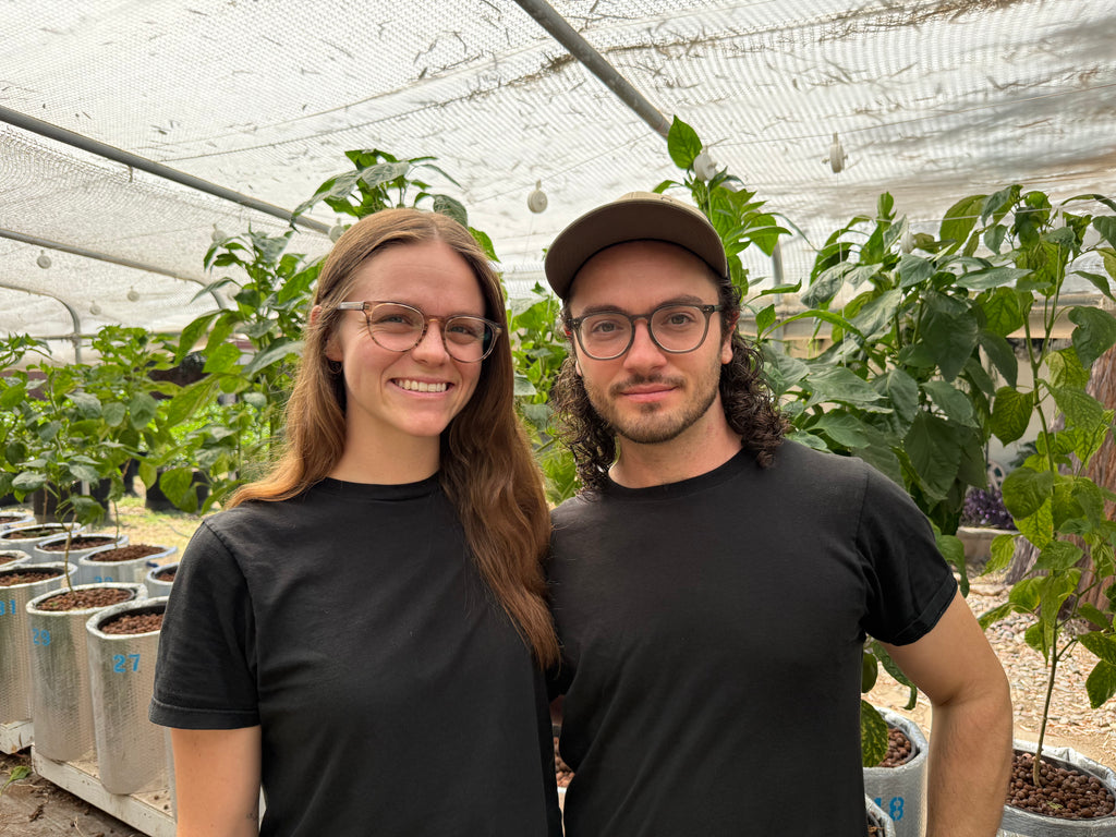 A smiling woman with glasses and a man wearing a cap and glasses stand together in a greenhouse, both wearing black t-shirts. Rows of potted plants are visible in the background under a white mesh canopy.